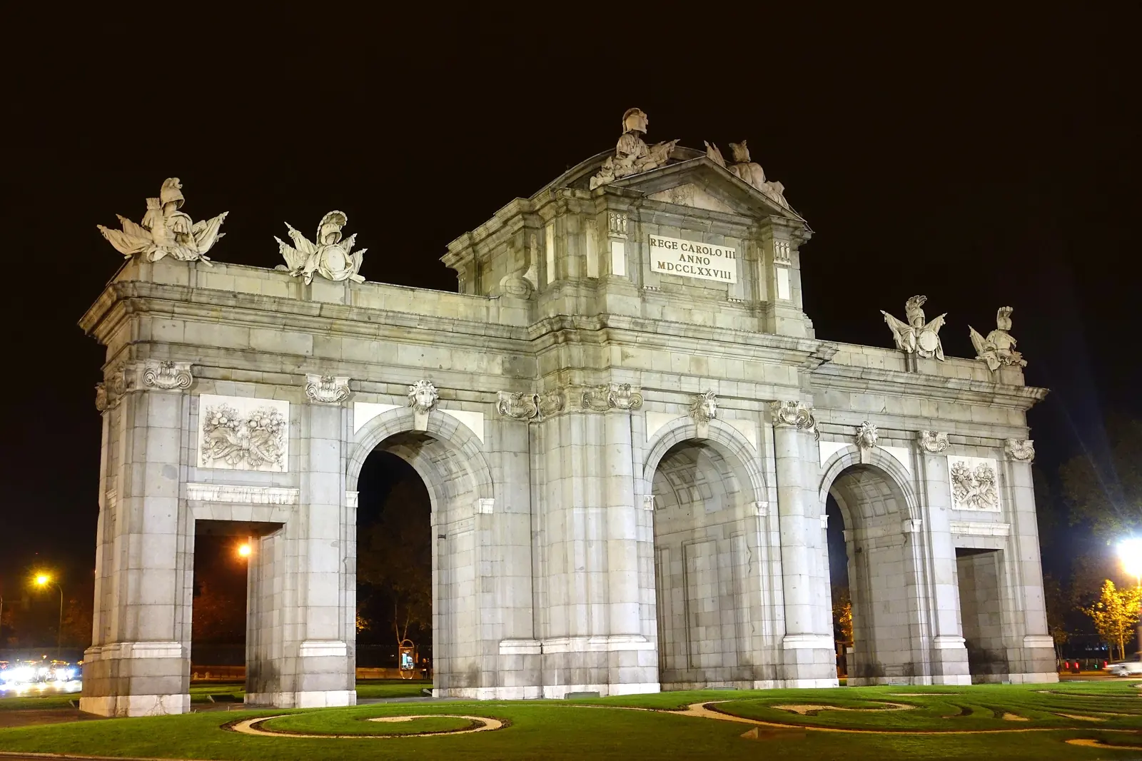 La Puerta de Alcalá iluminada de noche, una puerta de piedra pálida con tres arcos y figuras esculpidas en lo alto, situada en una glorieta ajardinada