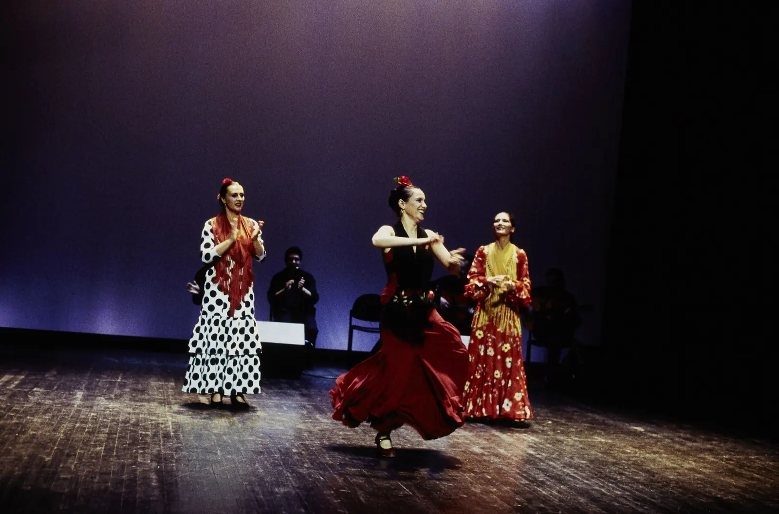 Tři flamenkoví tanečníci v šatech s puntíky a v červených šatech na tmavém jevišti ve fialovém světle, za nimi sedící hudebníci